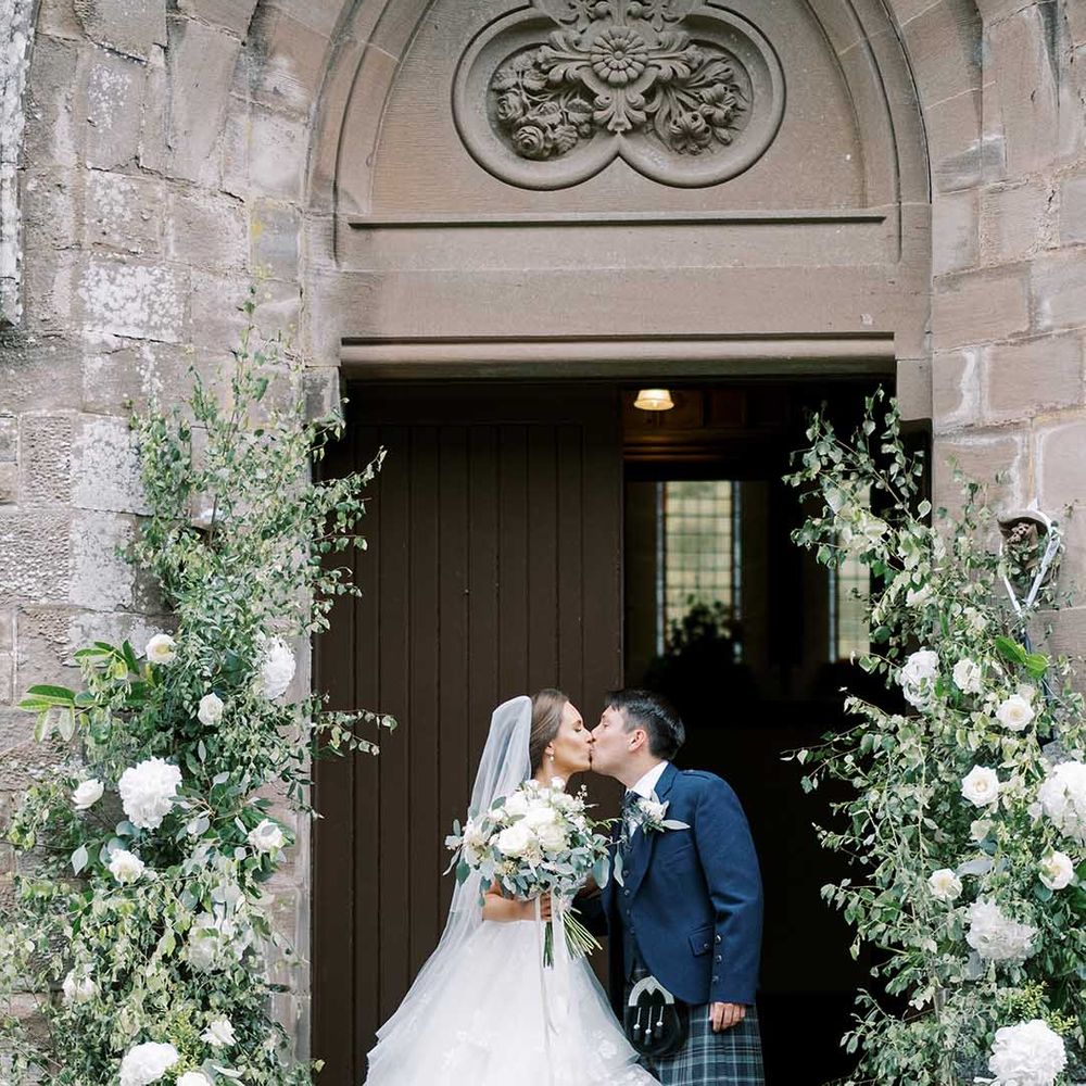 Bride in a Martina Liana princess wedding dress and groom in a wool suit and tart kilt kissing outside the church 