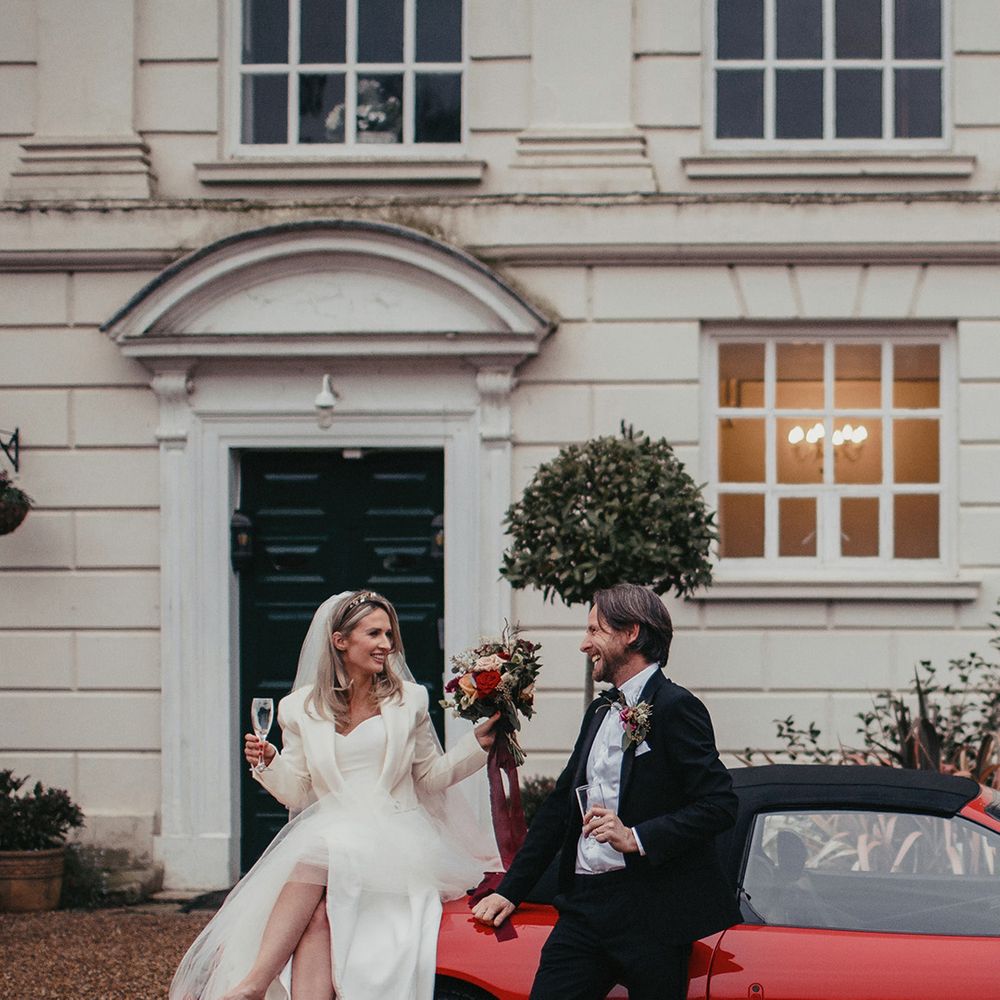 Bride & groom hold champagne glasses in front of red Ferrari at Gosfield Hall 