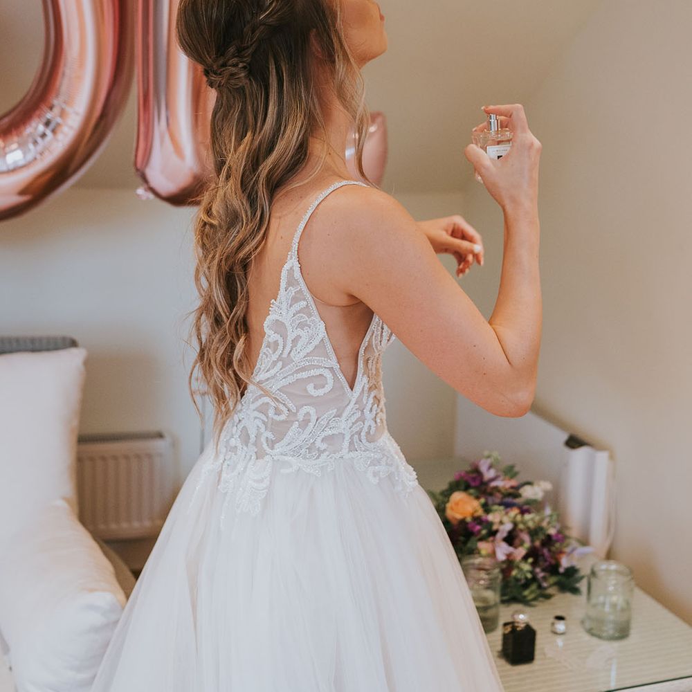 Bride puts on perfume in tulle dress with sparkly bodice and deep v cut back in front of rose gold balloons 