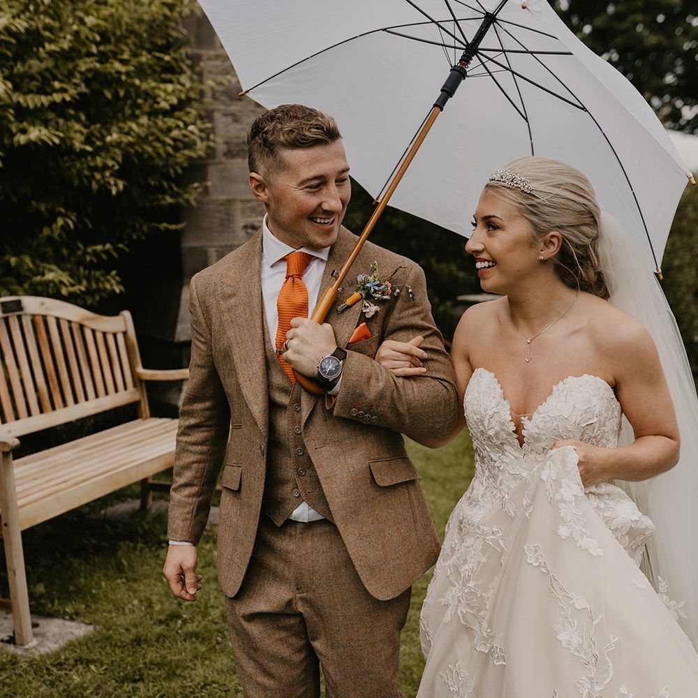 Groom in brown three piece suit with orange tie and handkerchief and colourful boutonnière with white umbrella and bride in sweetheart gown 