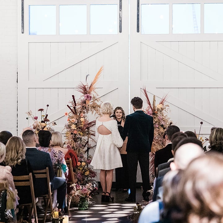 Bride and groom stand at the altar in front of colourful flower columns as guests watch on 