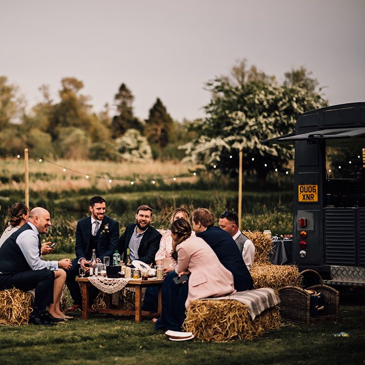 Wedding guests sit on hay bale seats as they talk around a table 