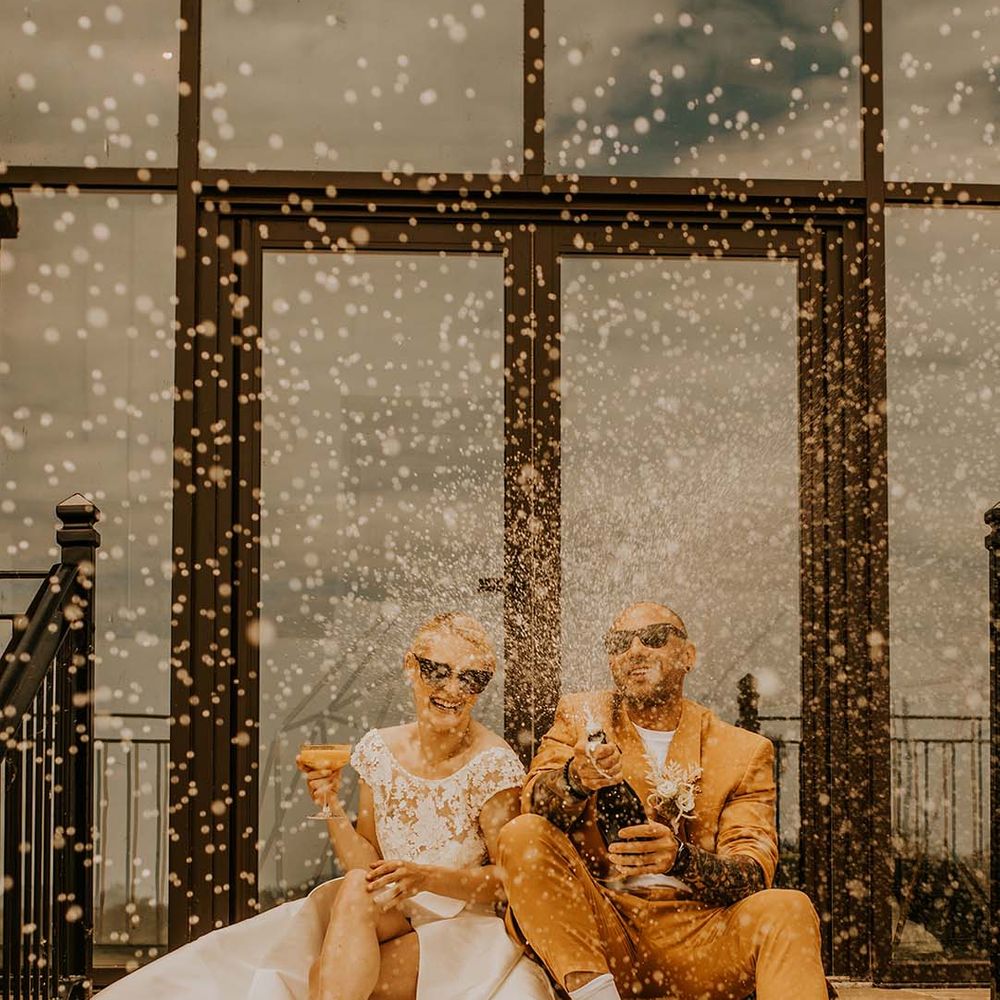 Bride in short sleeved wedding dress with gold heels and groom in mustard suit doing champagne pop