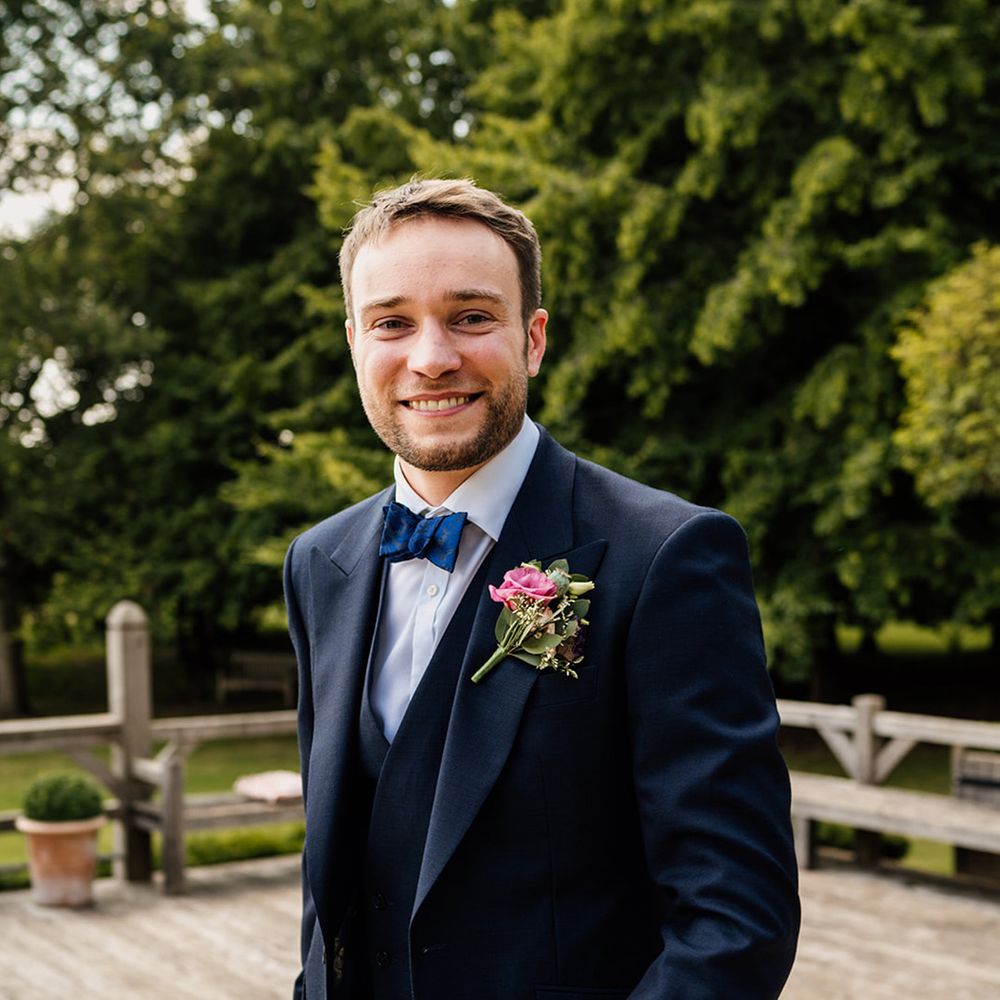 Groom in dark blue suit with blue bow tie with pink rose buttonhole for wedding 
