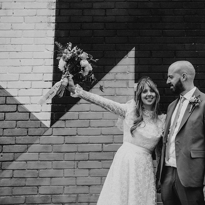 Bride wears vintage lace wedding dress with bow in her hair whilst standing beside groom who wears high street suit 
