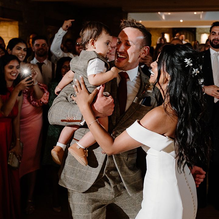 Bride with sparkly hair accessories in a fitted wedding dress with the groom in a brown suit dancing with their young son 