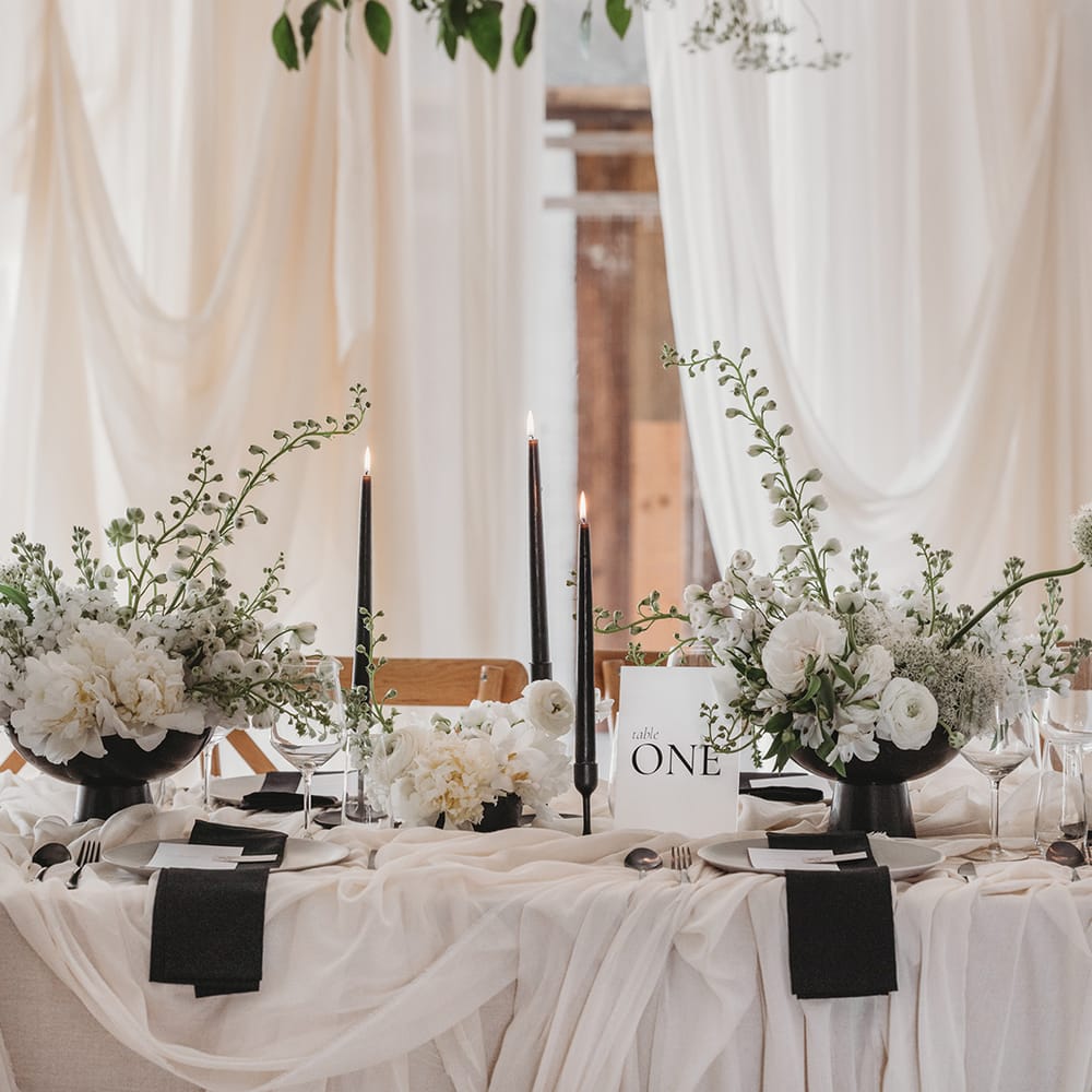Wedding tablescape with black taper candles, frosted acrylic table number sign, classic black and white stationery and white drapes 
