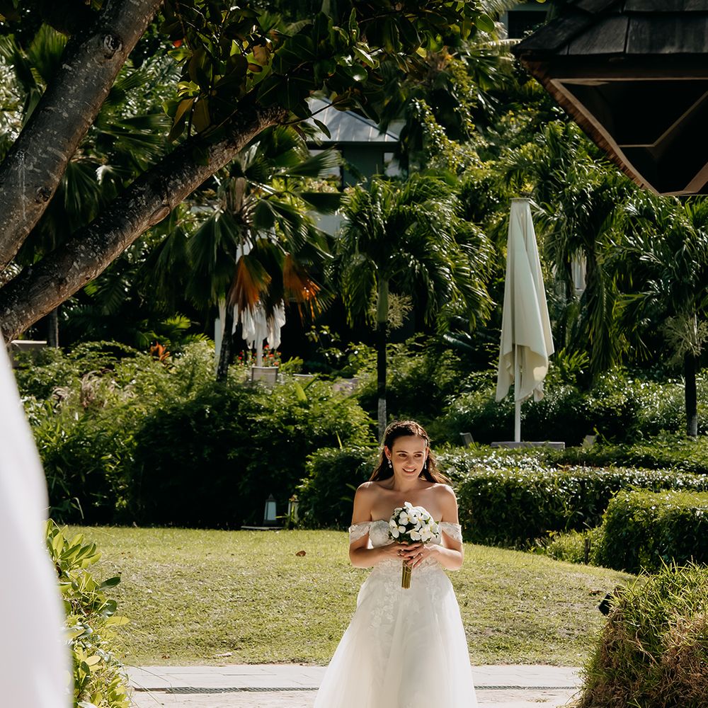 Bride walking on the grounds of Raffles hotel in Seychelles wearing off the shoulder lace wedding dress with sweetheart neckline and lace floral details and cathedral length veil holding white tropical bouquet 
