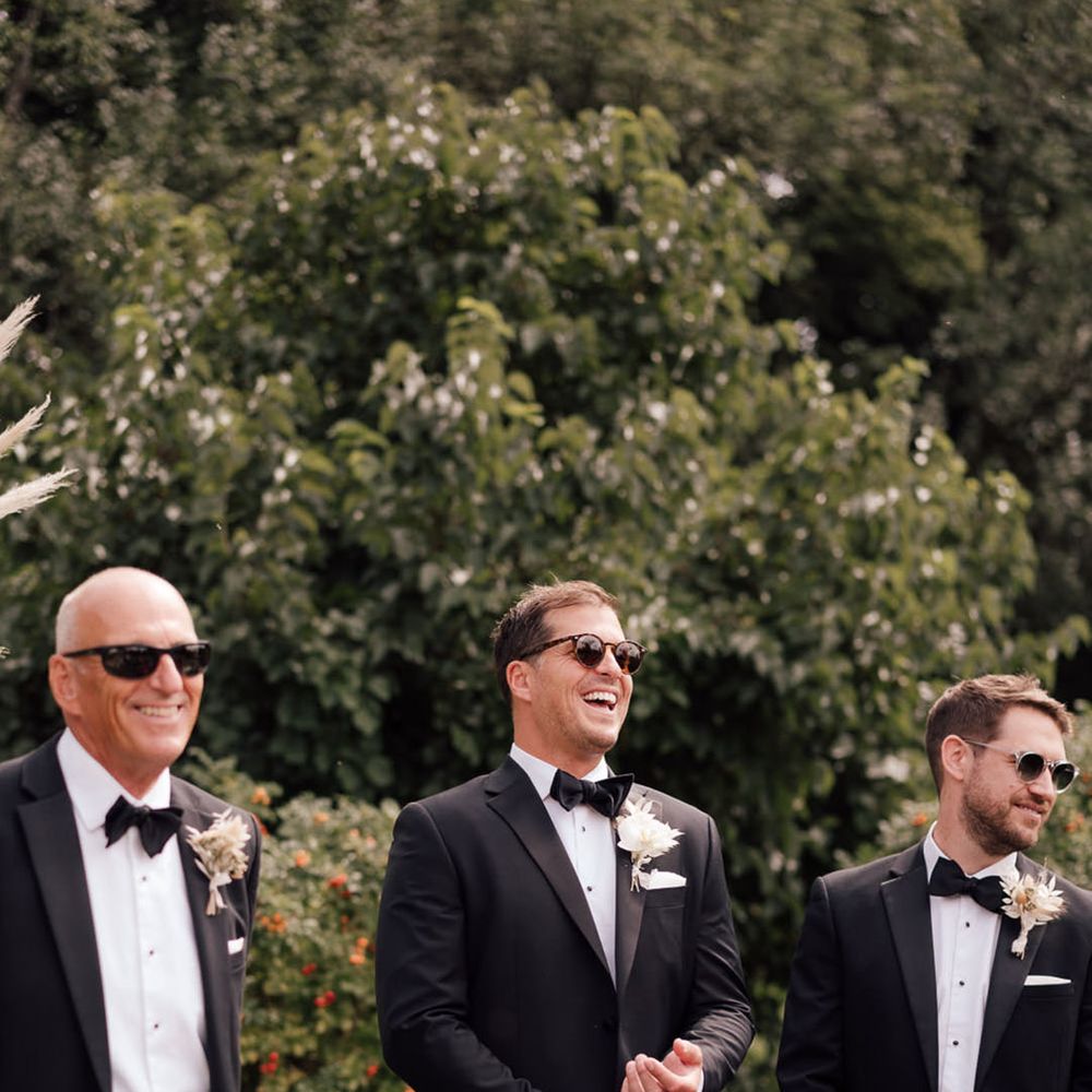 Groom and groomsmen stand together in black tie with sunglasses waiting at the aisle for the bride 