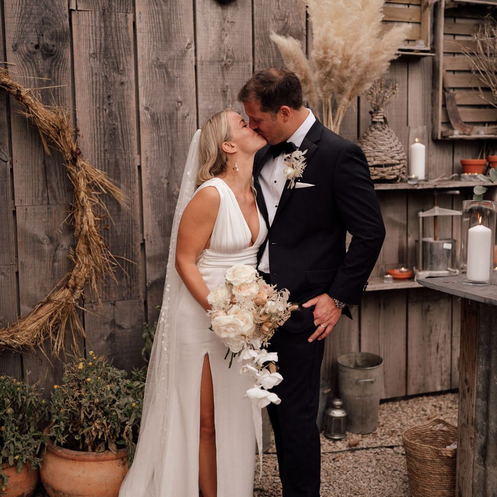 Bride in a satin white wedding dress with a cathedral length veil that has floral embroidery kissing the groom in a black tuxedo for boho luxe wedding 
