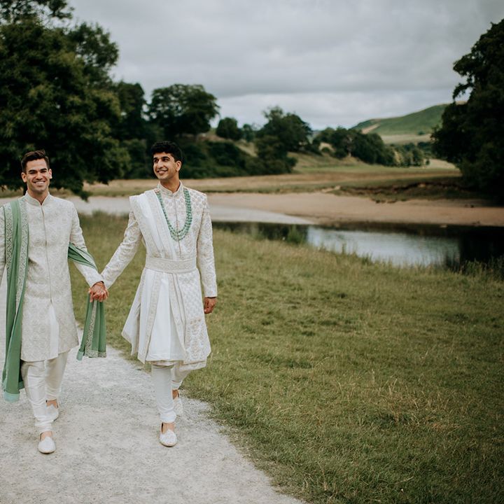 Grooms walk hand in hand wearing white and sage green sherwani's for couples portraits outdoors 