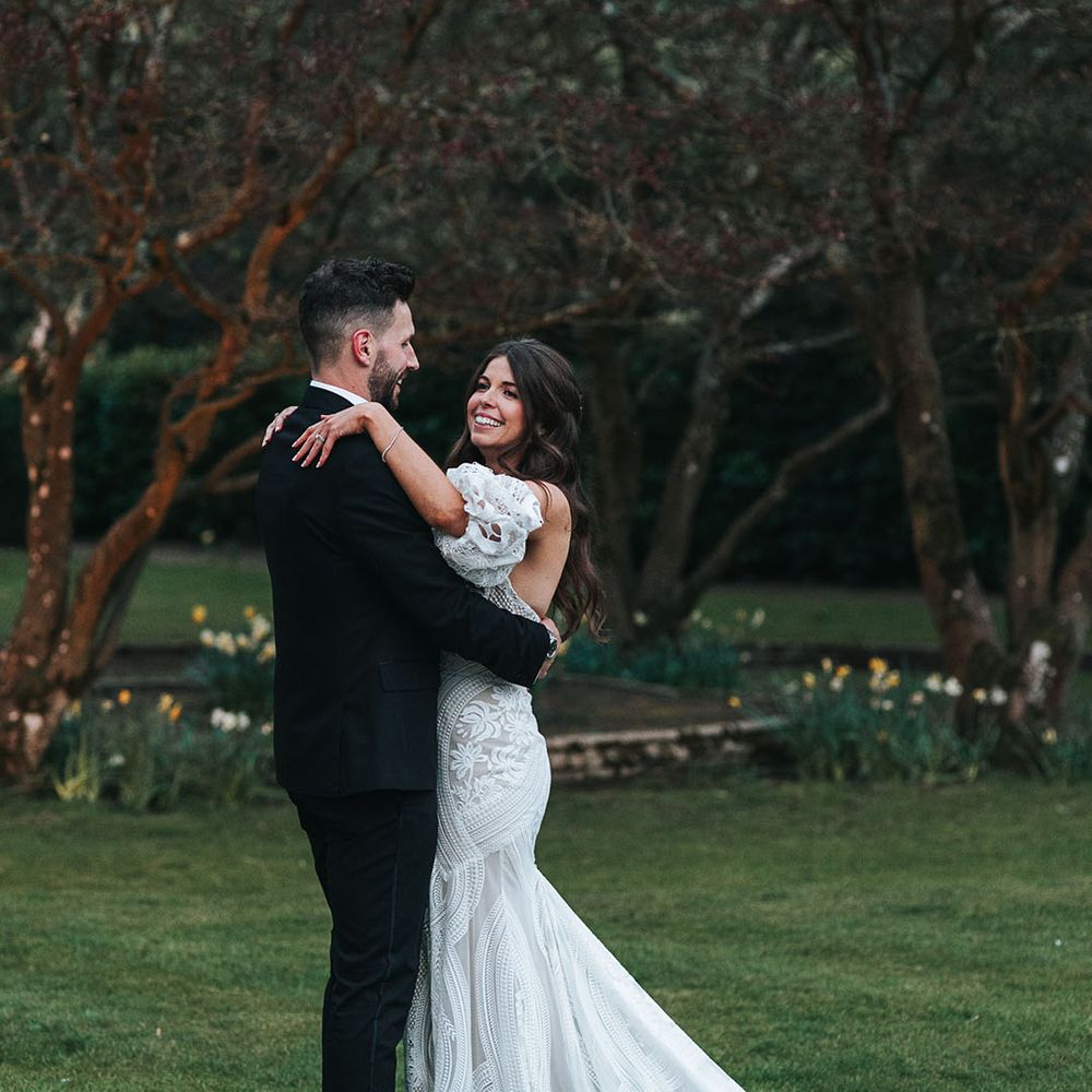 Boho lace strapless wedding dress with detachable sleeves sharing an intimate embrace with the groom in black tie 