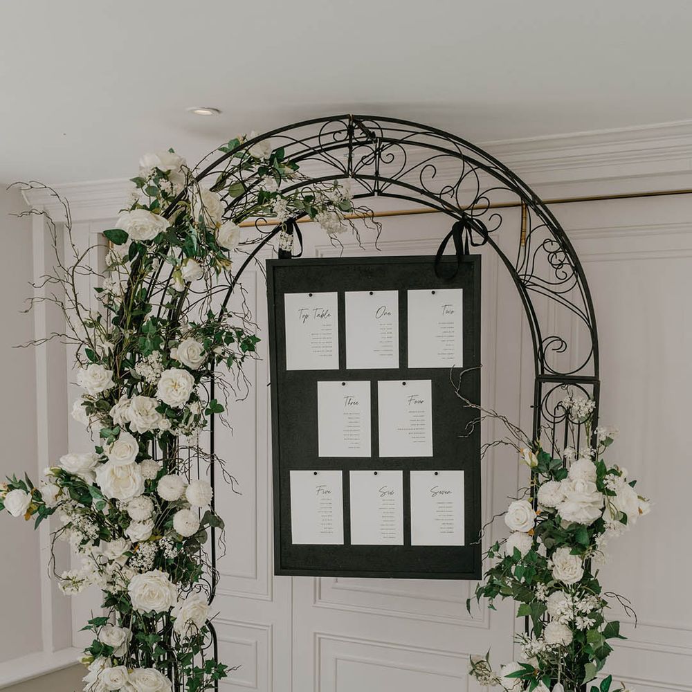 Black metal arch with black and white wedding seating chart with white wedding flowers 