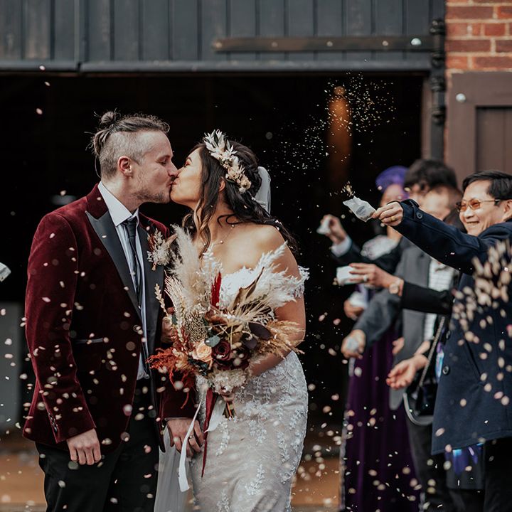 The groom in a velvet red suit jacket kissing the bride in a lace Zavana bridal wedding dress for their confetti exit 