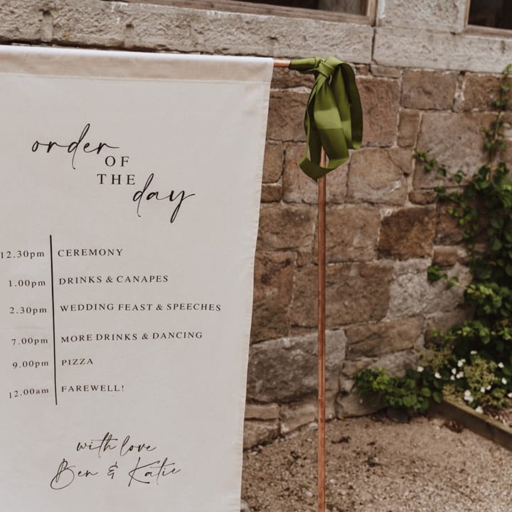 Black and white fabric order of the day wedding sign on a copper metal frame with green satin ribbon tied on the edges 