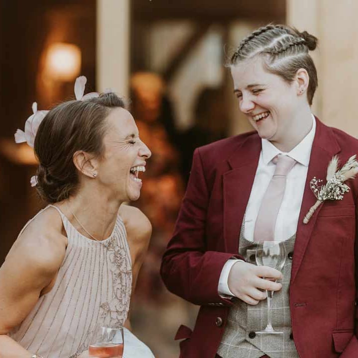 Bride in maroon bridal suit with grey waistcoat, dusky pink tie and autumnal dried flower boutonniere laughing with the mother of the bride in light pink sleeveless dress 