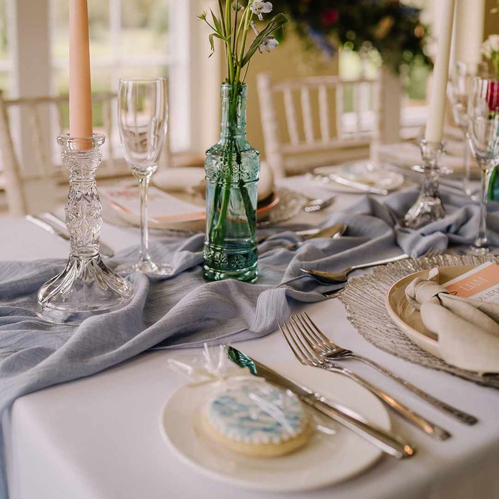 Blue glass bottle wedding centrepiece with dried spring florals, tapered candle in glass candlestick holder and cornflower blue wedding table runner 