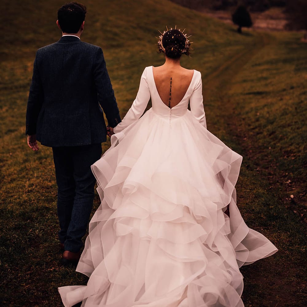 Bride in long sleeve ruffle gown with low back walking with the groom in navy suit 