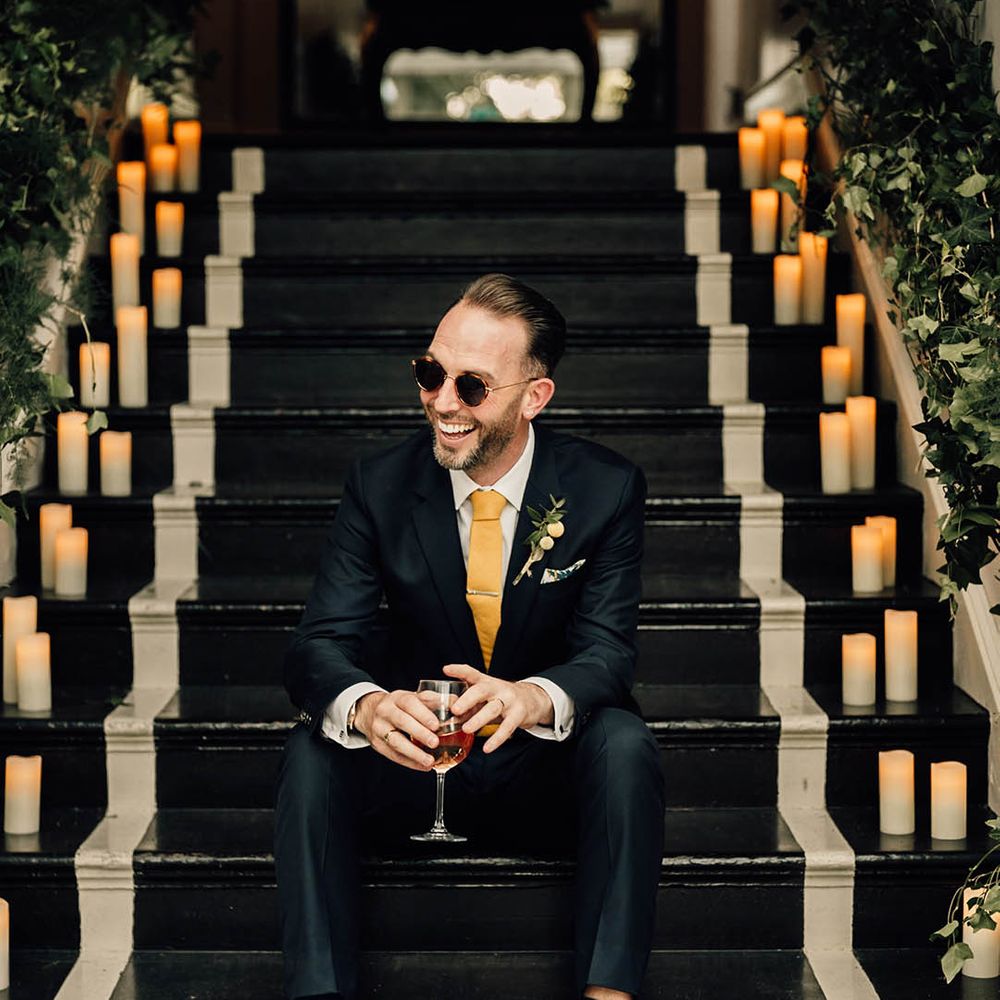 Groom in navy suit with yellow tie with yellow buttonhole and sunglasses groom accessories drinking glass of wine 