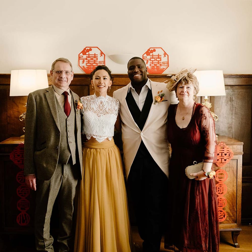 Groom incream morning suit with forest green grooms waistcoat and trousers, forest green pocket square and orange boutonniere standing with bride in white mesh lace bridal crop top and yellow wedding skirt and wedding guests during Chinese Tea Ceremony at Broadfield Court