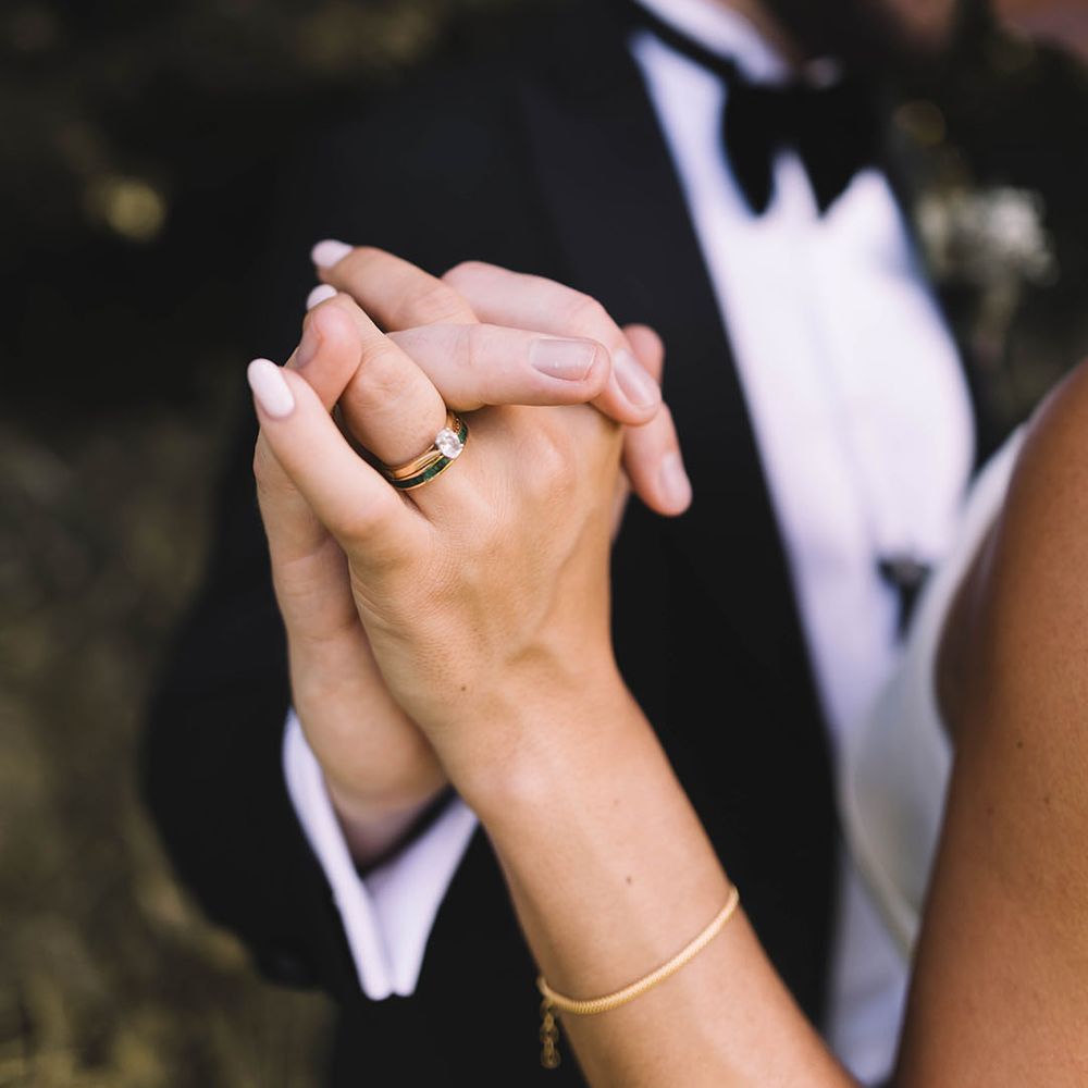 Bride wearing oval diamond engagement ring and gold wedding band in black tuxedo 