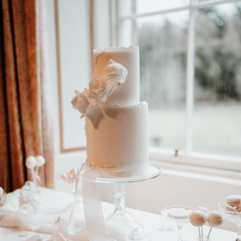 Two tier white iced wedding cake decorated with a white ribbon and white roses on glass cake stand with white cake pops, iced lollies and macarons