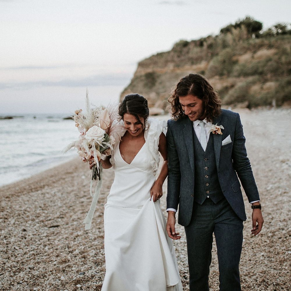 Groom in a navy blue suit walking on the beach with his bride in a Rime Arodaky ruffle sleeve wedding dress holding an orchid wedding bouquet 