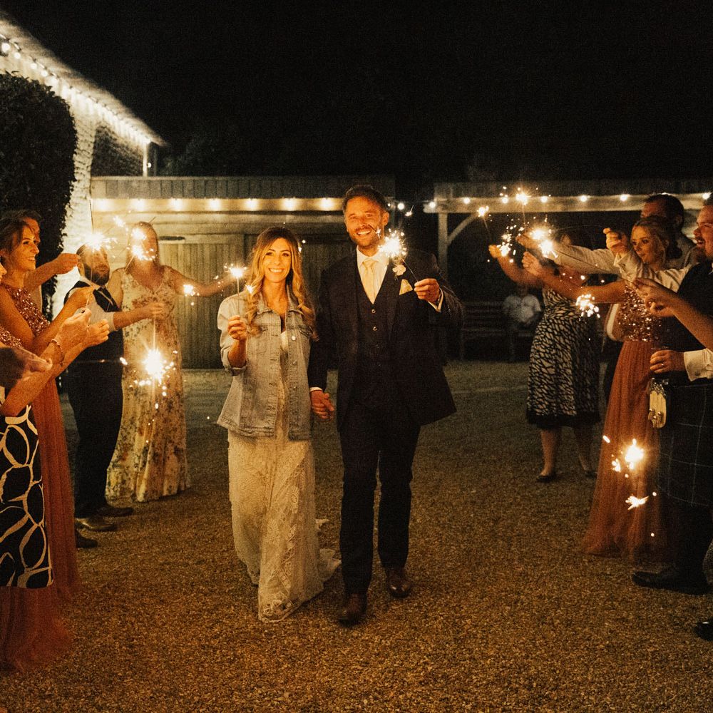 Bride in personalised denim jacket and groom hold sparklers with guests outside Tythe Barn Wedding venue