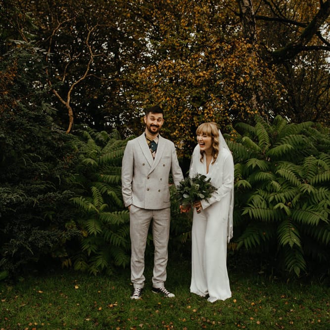 Portrait of the groom in a grey suit with patterned shirt and the bride in a jumpsuit with appliqué veil 