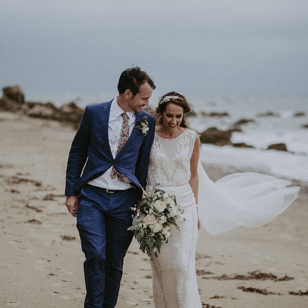Bride & groom walk along the beach together with the sea in the background