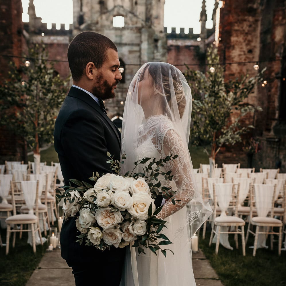 Bride and groom embracing at the altar with the bride wearing a sheer veil over her face and holding a white rose wedding bouquet 