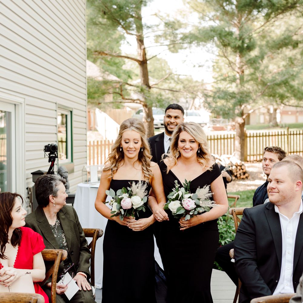Two bridesmaids in black bridesmaids dresses and curled hair walk down the aisle holding white, pink and green rose wedding bouquets at backyard wedding