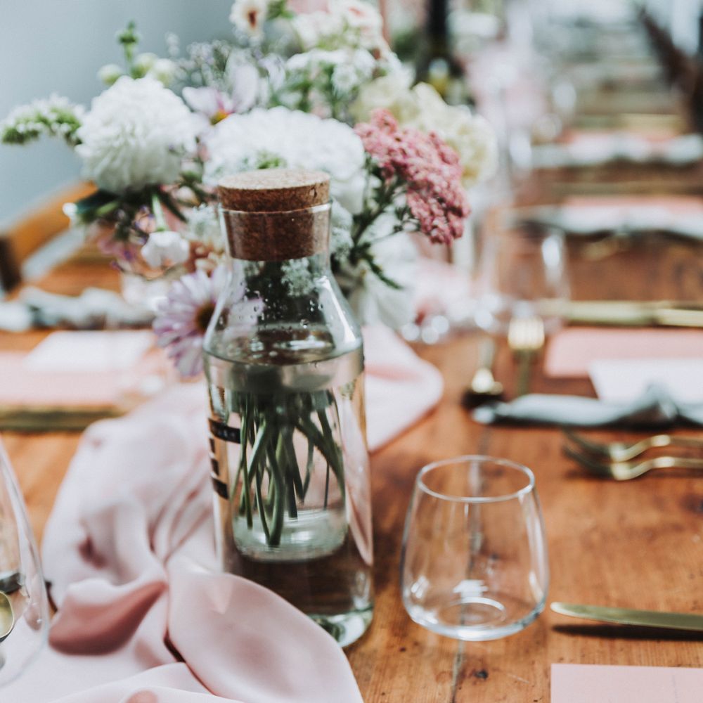Pink table cloth runner across wooden table complete with glass water bottles with cork tops and gold cutlery 