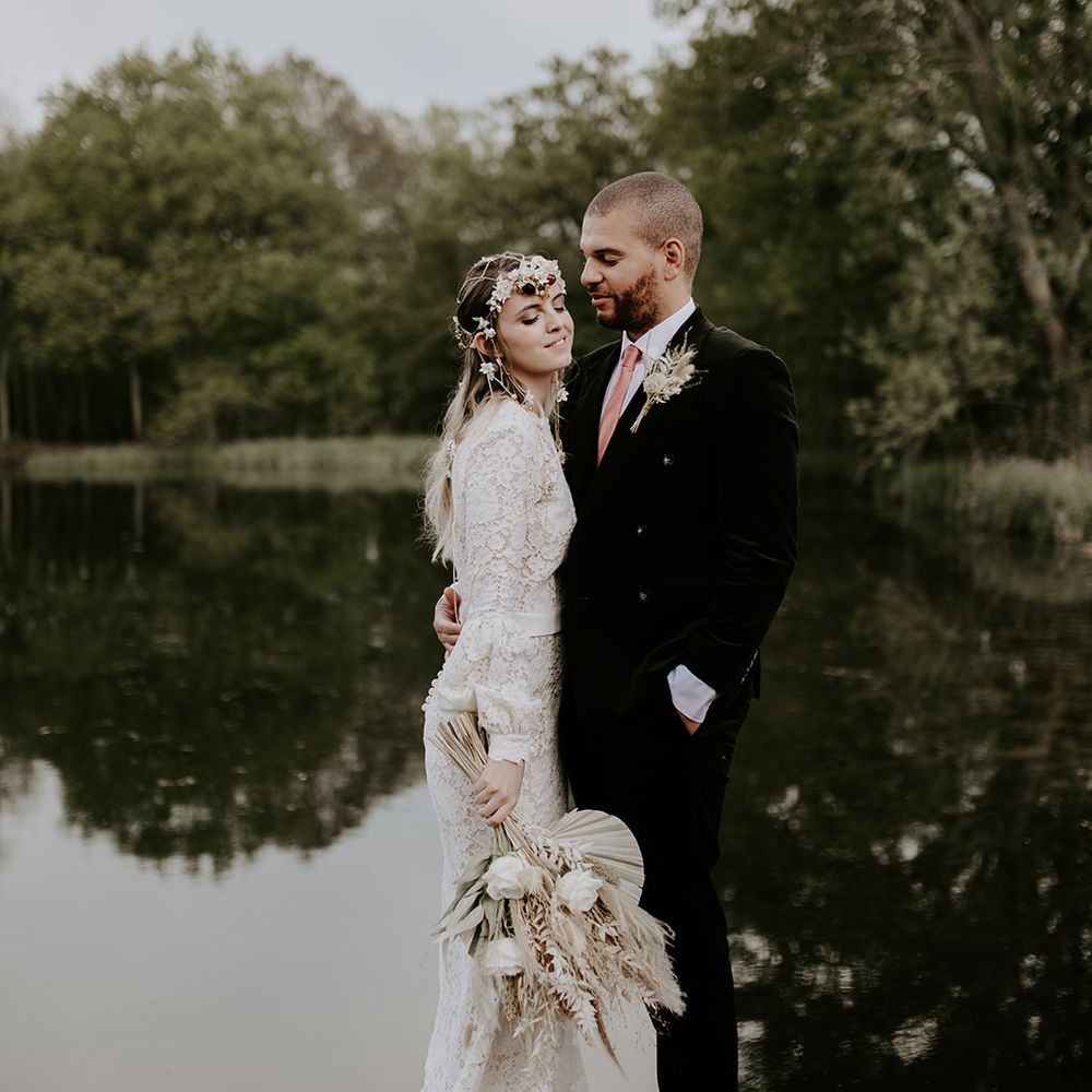 Boho bride in a lace wedding dress with long sleeves and puddle train holding a dried flower bouquet embracing her husband on the dock at Waresley Park Estate 