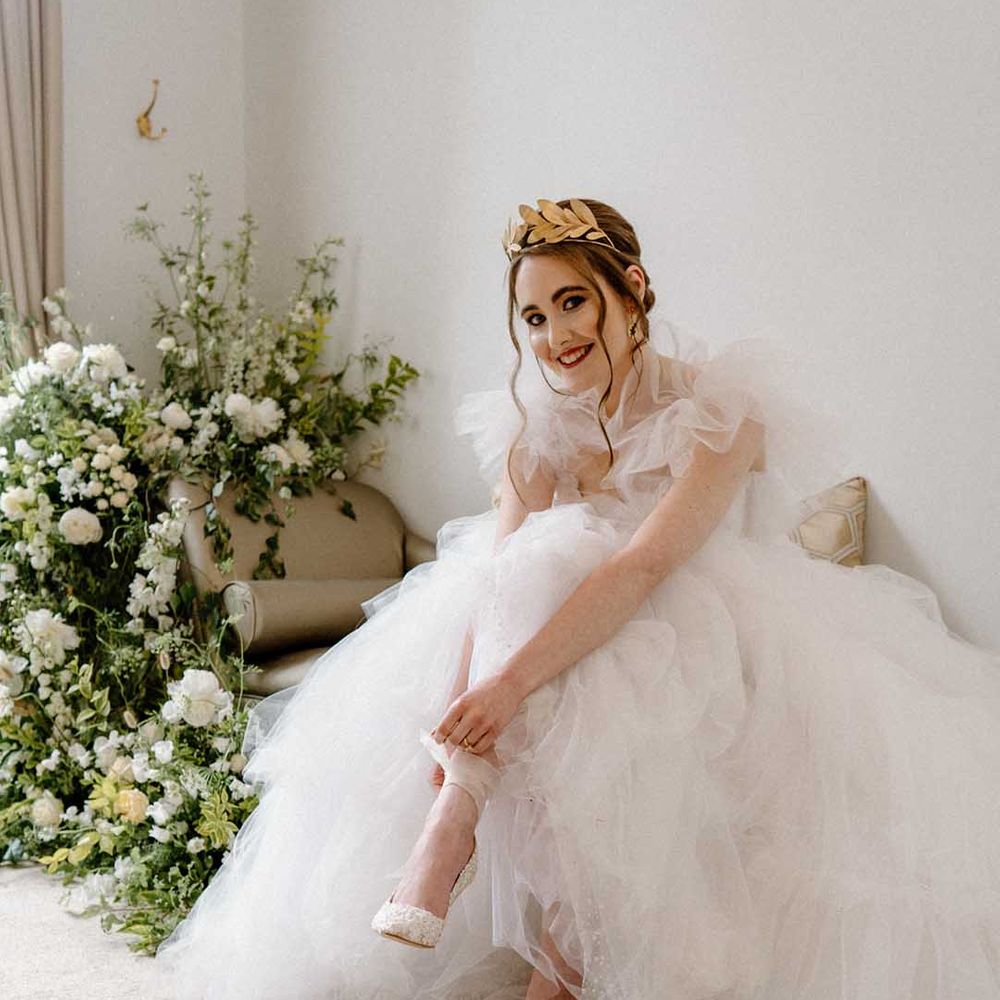 Bride in a tulle wedding dress with gold crown sitting on a chaise lounge putting on her embellished bridal shoes with ribbon ankle straps 