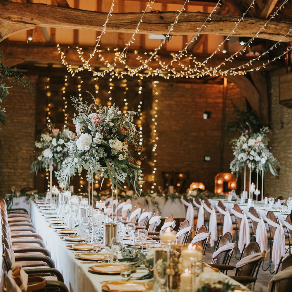 White, pink and green flower bombs on stands at long wedding breakfast table lit with strung fairy lights at Tythe Barn wedding with barn wedding flowers