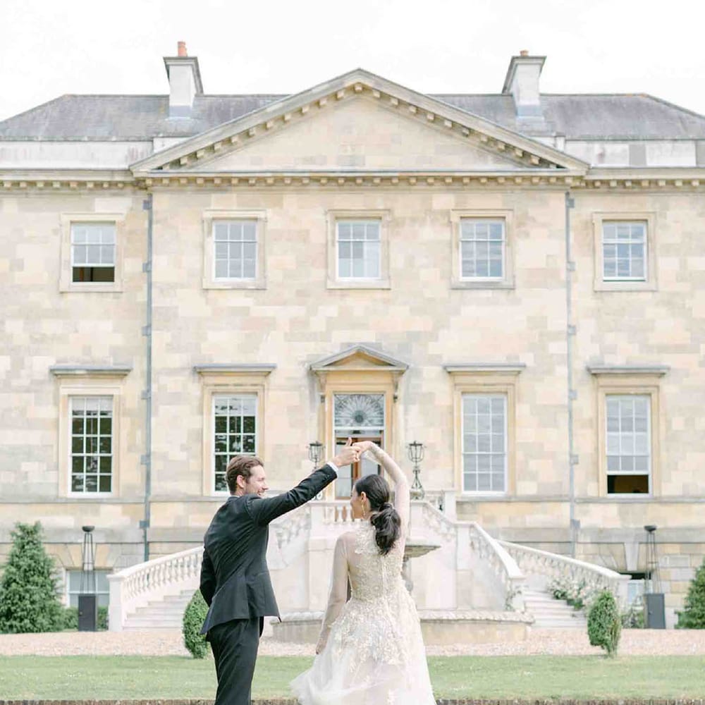 Groom in a tuxedo twirling his bride in a vintage lace wedding dress with long sleeves on the grounds of Botley's Mansion, Surrey 