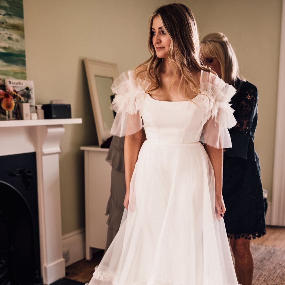 Bride in white Halfpenny London Mayfair dress with ruffle sleeves gets her dress zipped up by Wedding guests before church ceremony in Cornwall