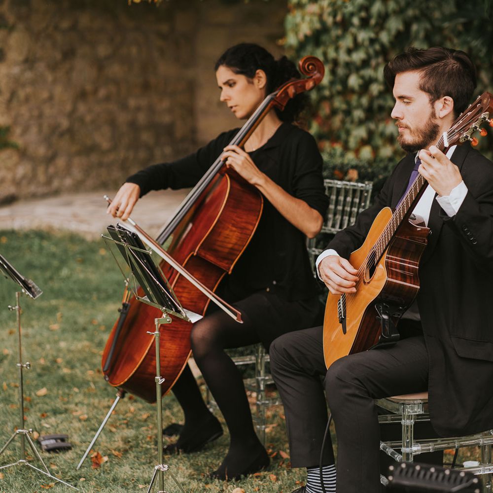 Guitar and cello player sit in perspex bamboo chairs playing outside during outdoor wedding ceremony at Notley Abbey