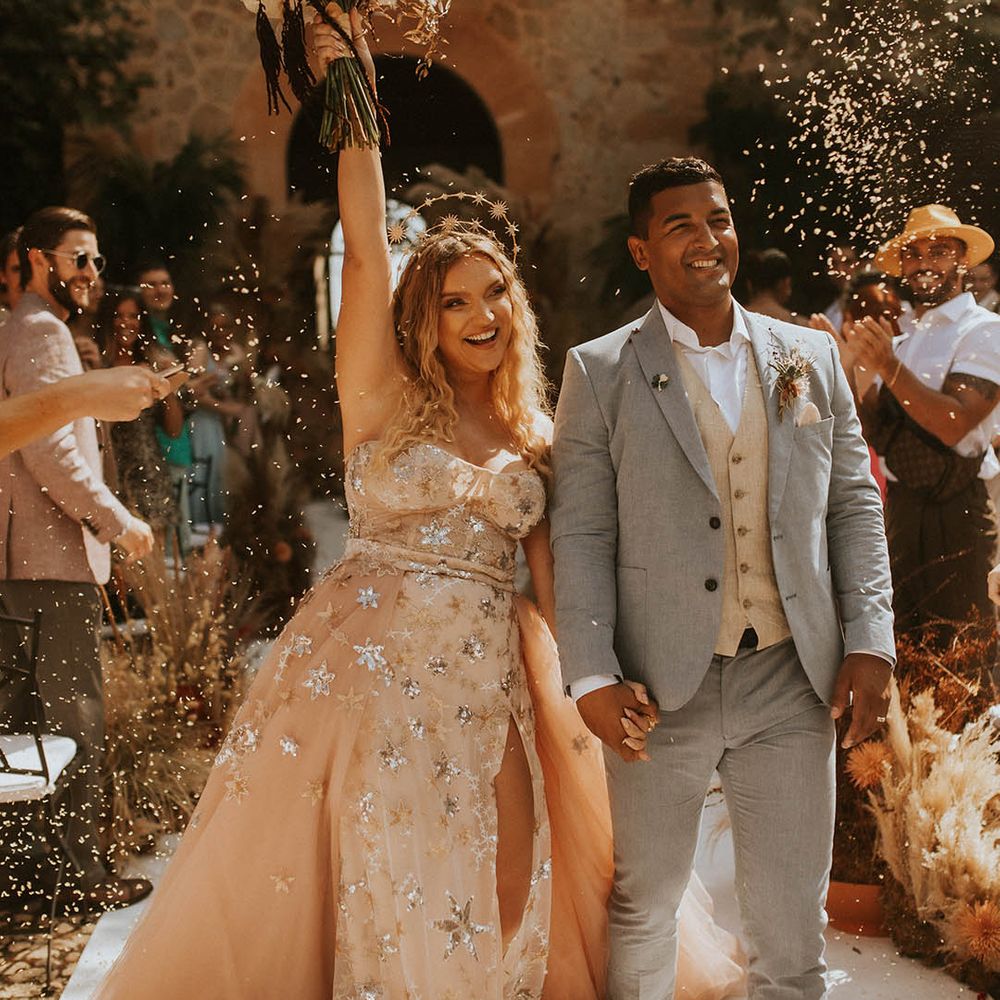 Bride & groom walk through confetti on their wedding day as bride lifts her bouquet in celebration