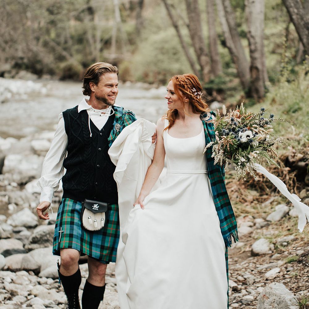 highland wedding inspiration with groom in a tartan kilt and bride in a princess wedding dress with plaid sash walking through a stream 
