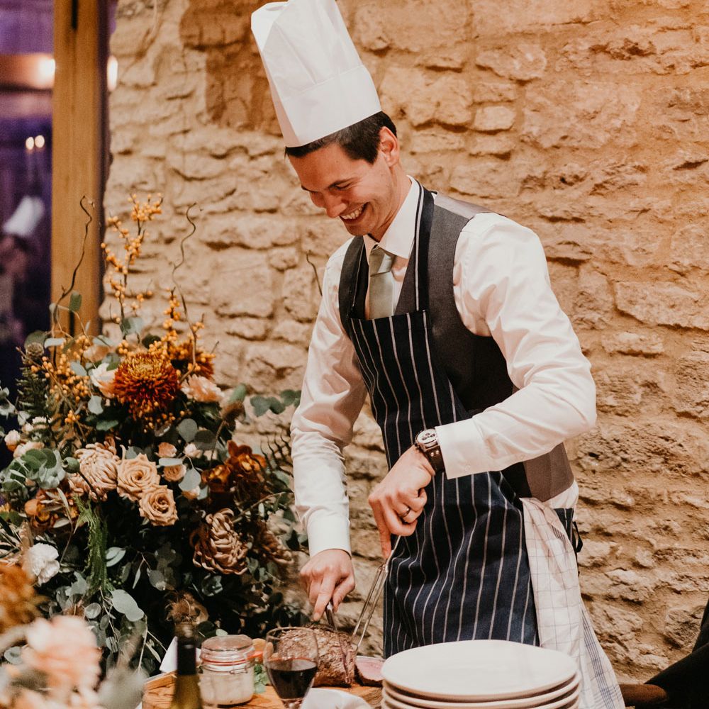 Smiling groom in striped apron and chefs hat cuts up meat at table during rustic barn wedding breakfast 