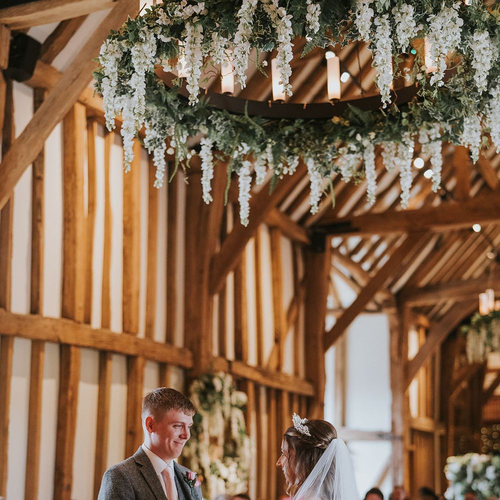Groom in grey tweed suit and pink tie with bride at the altar with large rustic chandelier decorated with hanging wisteria 