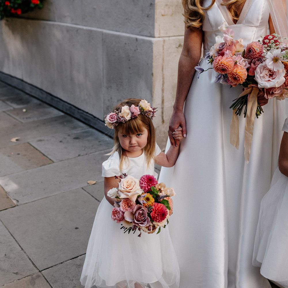 Cute flower girl in a white dress and flower crown holding a blush pink bouquet with roses and dahlias 