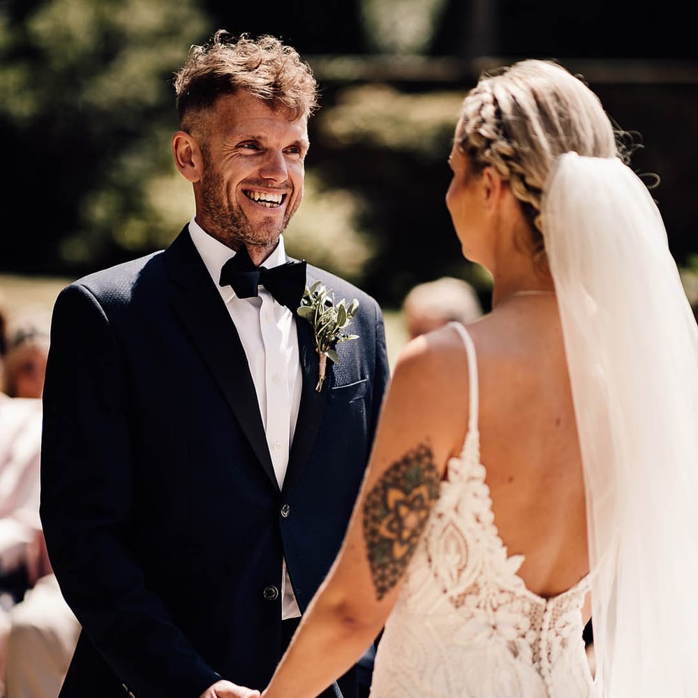 Groom in blue tuxedo and black bow tie smiles as he looks at the bride and they hold hands together for outdoor ceremony 