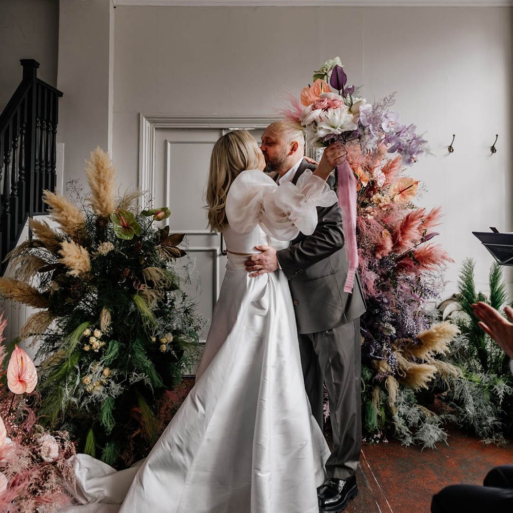 Groom kissing his bride in a JESUS PEIRO wedding dress at their wedding ceremony surrounded by colourful colour flower arrangements 
