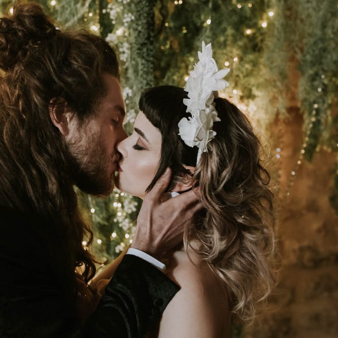 Groom with long hair kissing his bride in a white 3D flower headband at altar with a twig, fairy light and foliage backdrop 
