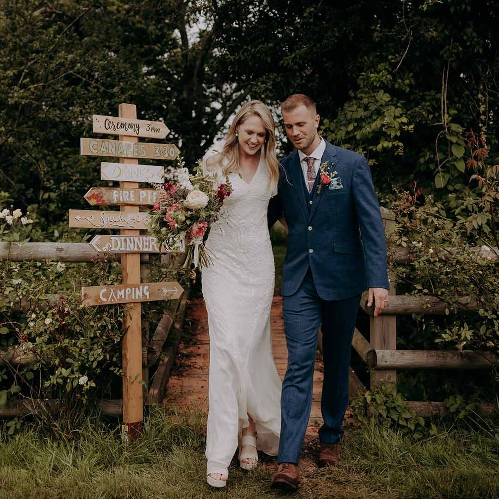Bride in short sleeved embellished wedding dress and groom in dark suit standing by DIY wedding sign