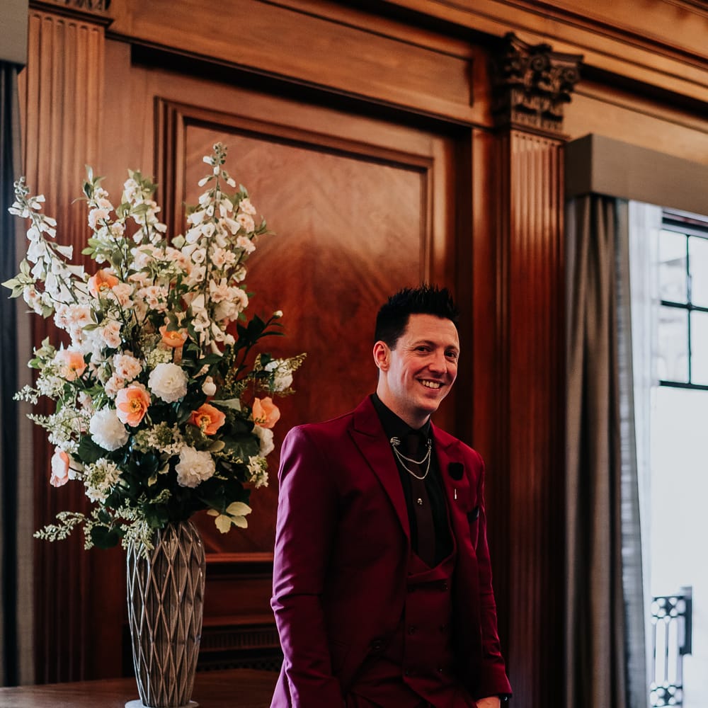 Groom wears wine red suit and black shirt complete with chain detailing 
