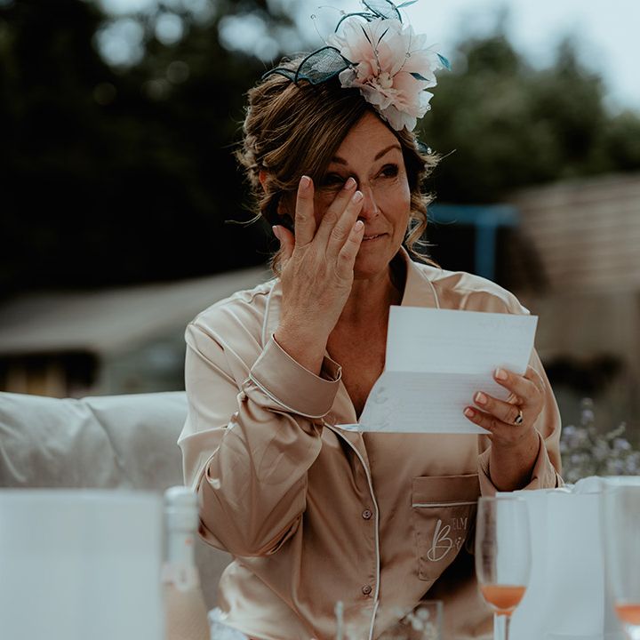 Mother of the bride in gold satin pyjamas wears a pink and green fascinator and wipes her eyes as she reads an emotional letter